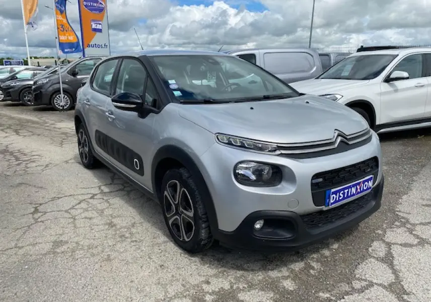 Citroën C3 gris clair vue 3/4 avant droit avec enjoliveurs 3D et protections latérales noires sur parking.