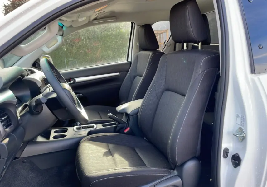 Intérieur du Toyota Hilux 2025 blanc, vue côté gauche sur les sièges avant en tissu noir et tableau de bord.