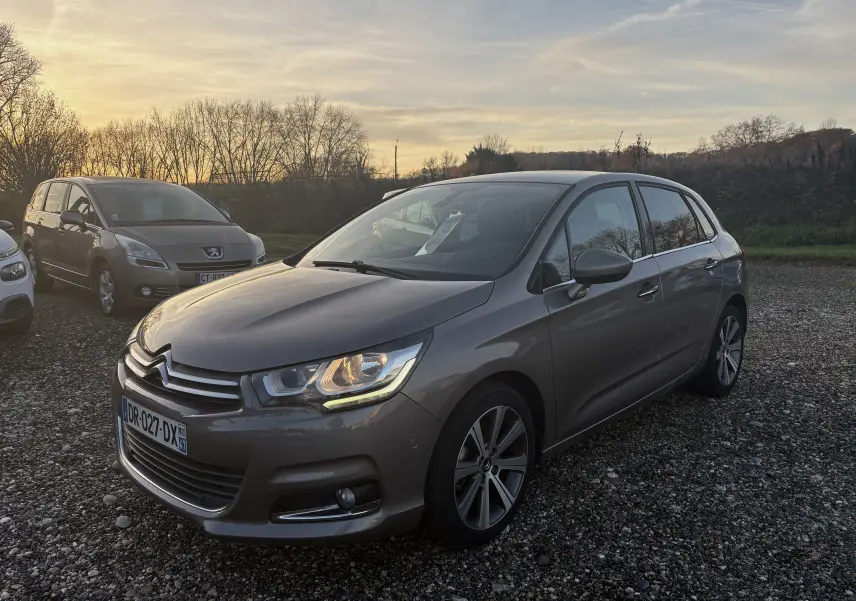 Citroën C4 II gris métallisé vue 3/4 avant droit au coucher du soleil sur un parking gravillonné.