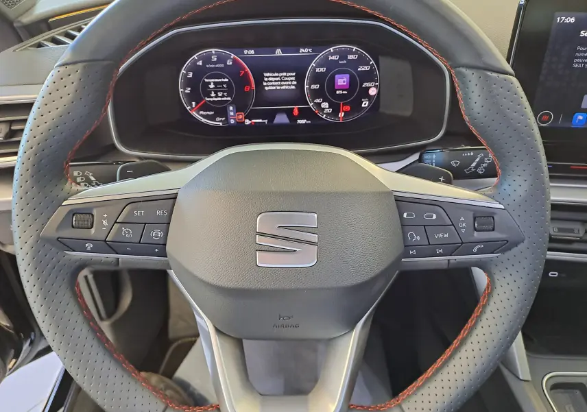 Volant perforé avec surpiqûres rouges de la SEAT LEON 1.5 eTSI Hybrid 2024, tableau de bord numérique visible en arrière-plan.