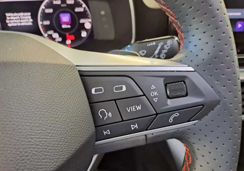 Gros plan sur les commandes au volant perforé noir avec surpiqûres rouges du SEAT LEON 1.5 eTSI Hybrid 2024.