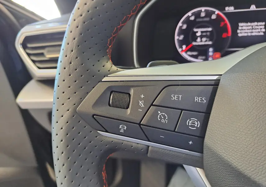 Gros plan sur les commandes au volant perforé avec surpiqûres rouges du SEAT LEON 1.5 eTSI Hybrid 2024, tableau de bord en arrière-plan.
