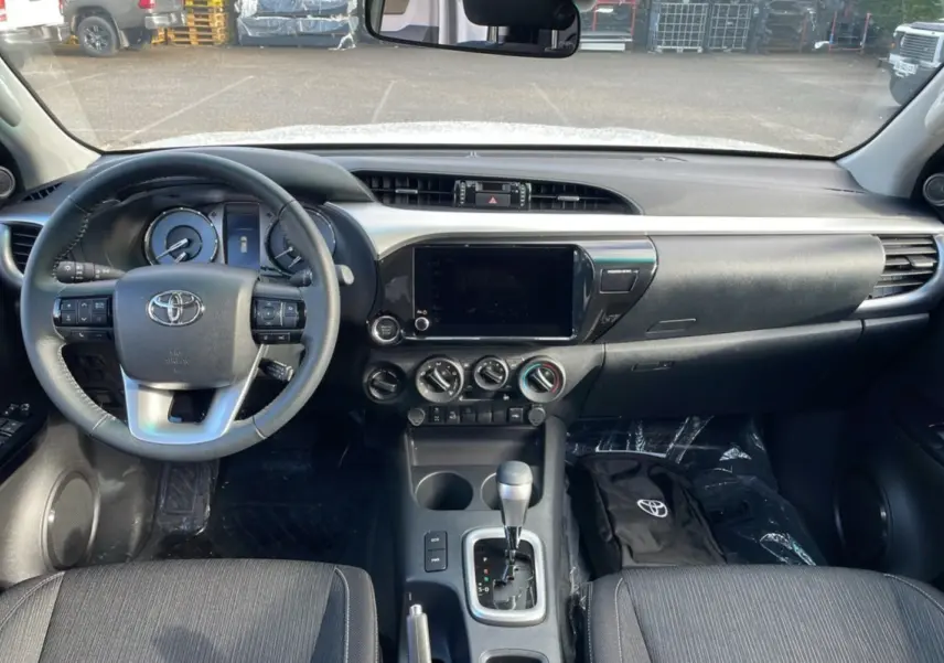 Intérieur du Toyota Hilux 2025 en vue frontale, tableau de bord noir avec écran tactile et levier de boîte automatique.