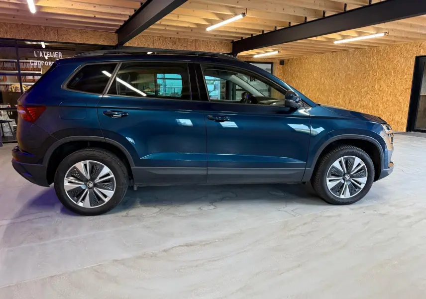 Vue de profil côté gauche d'un Skoda Karoq bleu 2023 avec jantes alu 17 pouces et toit noir dans un garage.