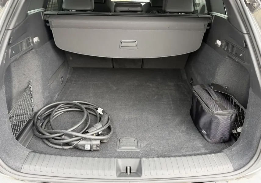 Coffre arrière ouvert de l’Audi Q6 e-tron gris, vue plongeante montrant câble de recharge et sac de rangement noir.