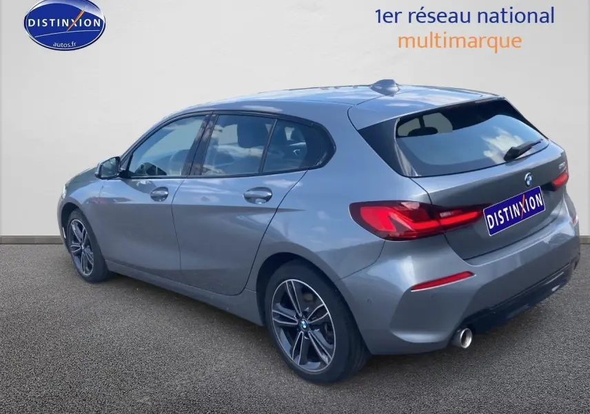 BMW Série 1 gris foncé vue 3/4 arrière côté gauche, avec jantes alliage et feux arrière LED distinctifs.
