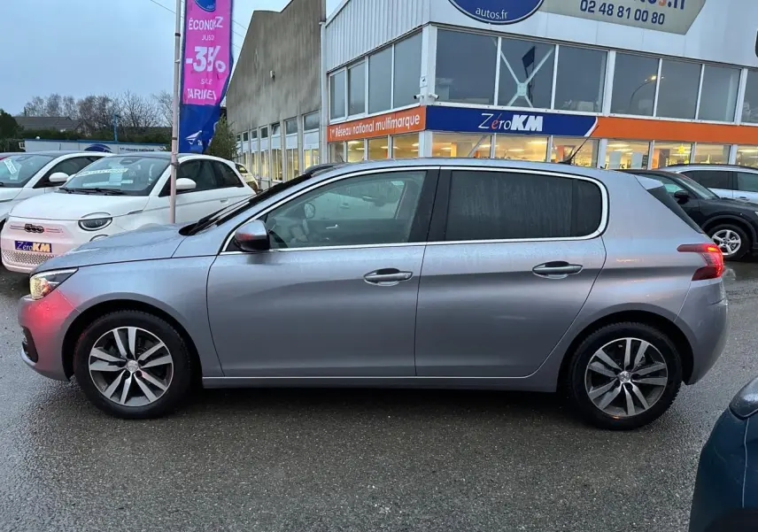 Profil côté gauche d'une Peugeot 308 gris Artense 2018 garée devant un concessionnaire sous la pluie.