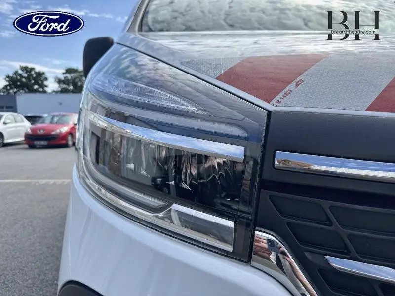 Gros plan sur le phare avant droit du Renault Trafic Fourgon blanc glacier, soulignant les détails du bloc optique et la calandre noire.