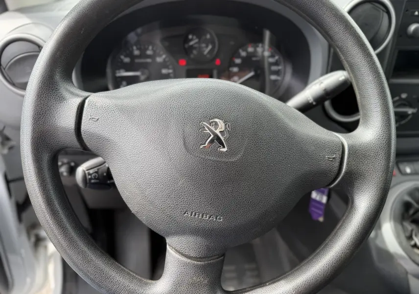 Gros plan sur le volant noir avec logo Peugeot du Partner blanc 2017, tableau de bord et commandes visibles en arrière-plan.