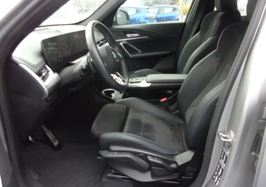 Vue intérieure côté conducteur du BMW X1 argent métallisé 2024, sièges sport noirs avec surpiqûres bleues et volant cuir.