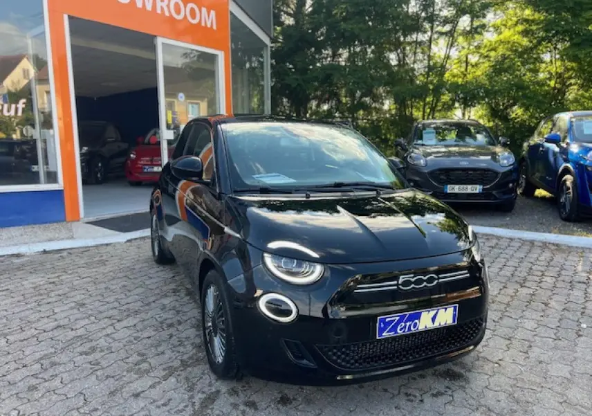 Vue 3/4 avant d'une FIAT 500 électrique noire stationnée devant un showroom avec phares LED ronds allumés.