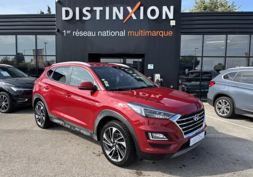 Hyundai Tucson rouge métallisé en 3/4 avant droit, avec jantes alliage et calandre chromée visible devant un bâtiment noir Distinxion.