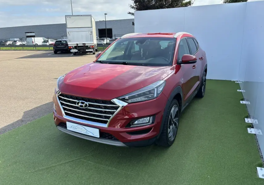 Vue 3/4 avant d'un Hyundai Tucson rouge métallisé garé sur un sol vert avec calandre chromée et jantes alliage.