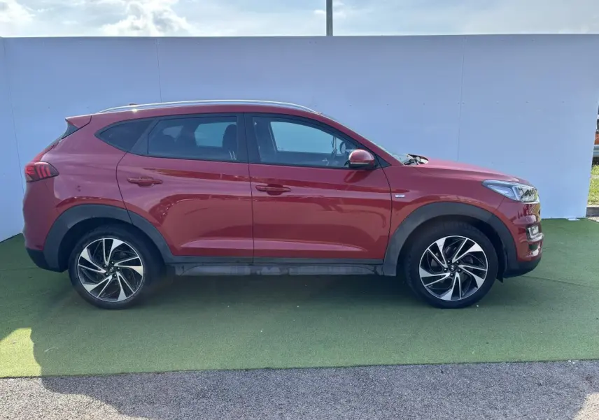 Profil droit d'un Hyundai Tucson rouge métallisé 2020 avec jantes alliage et vitres teintées sur fond neutre.