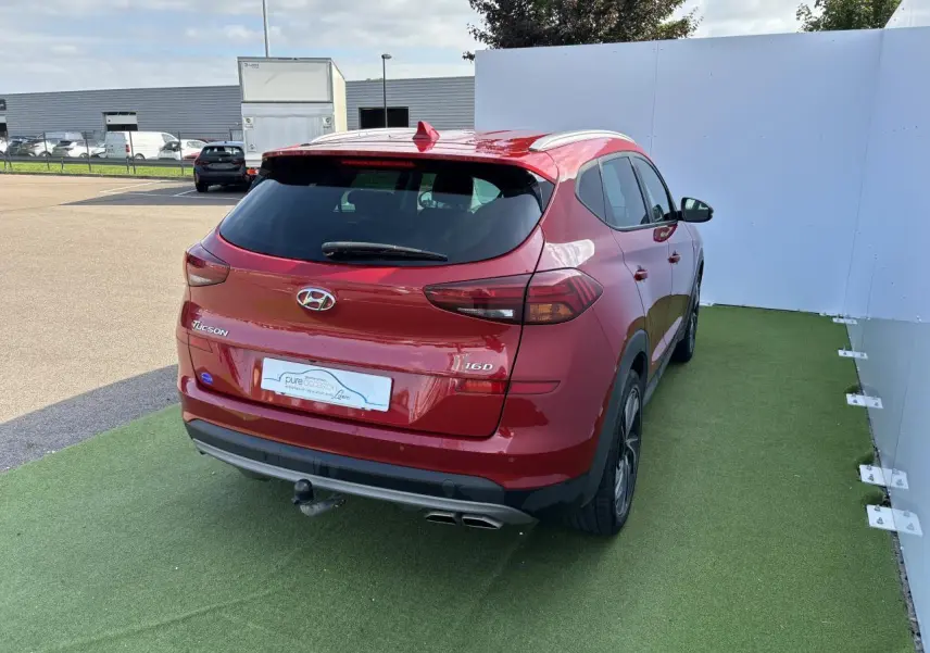 Vue 3/4 arrière droite d'un Hyundai Tucson rouge métallisé avec attelage et feux arrière LED visibles.