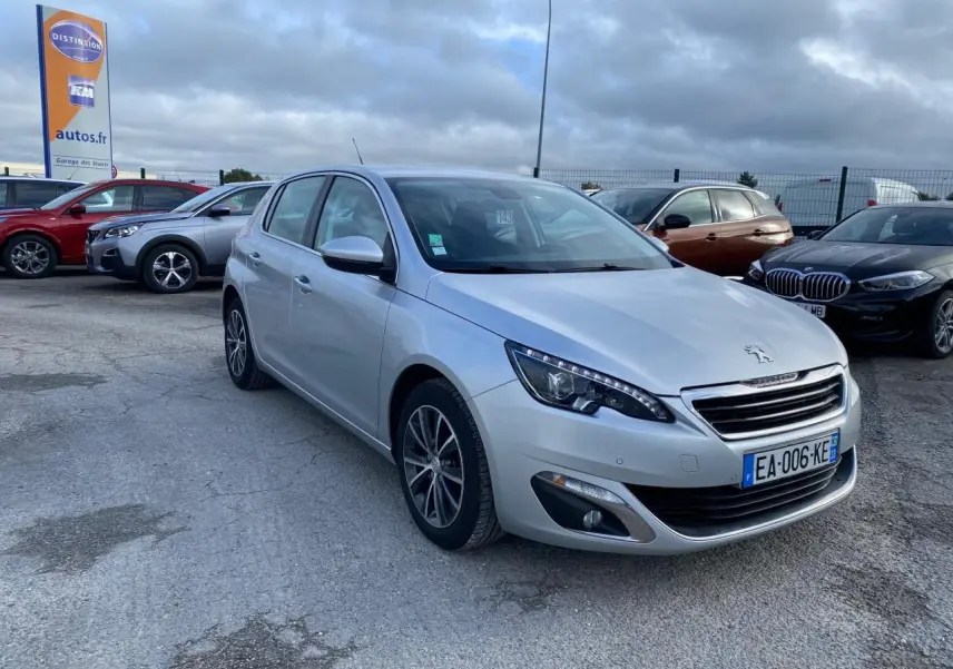 Peugeot 308 gris aluminium vue 3/4 avant droit, avec phares LED et jantes alliage sur parking extérieur.