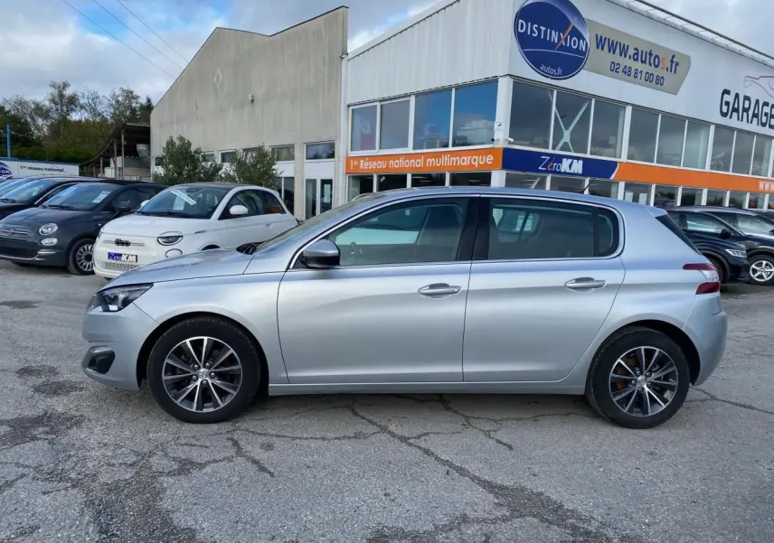 Profil côté gauche d'une Peugeot 308 gris aluminium 2016 garée devant un garage multimarque.