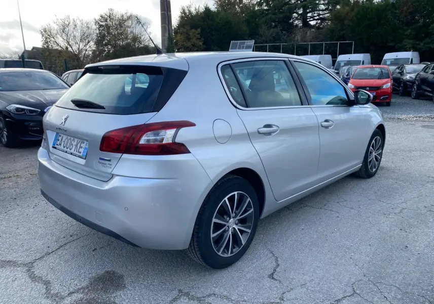 Vue 3/4 arrière droite d'une Peugeot 308 gris aluminium garée sur un parking avec plusieurs autres véhicules.
