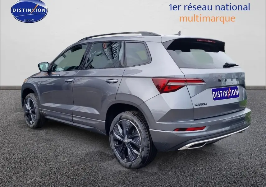 Vue 3/4 arrière droite d'un SKODA KAROQ gris graphite avec jantes alu et barres de toit visibles.