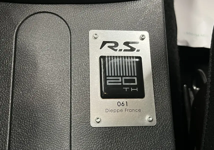 Plaque numérotée n°61 de la Renault Clio III RS 20th Anniversary sur fond noir texturé, vue rapprochée intérieure.