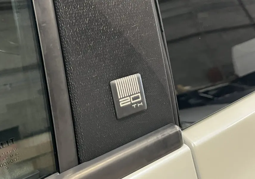 Gros plan sur l’écusson « 20TH » noir et blanc sur la monture de porte d’une Renault Clio III blanc nacré toit noir.