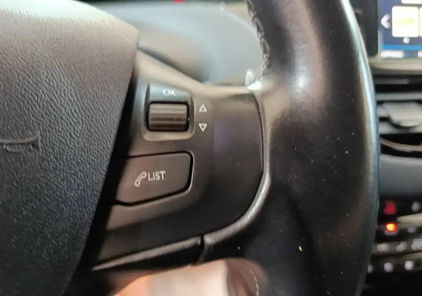 Gros plan sur le volant cuir noir de la Peugeot 2008 avec commandes vocales et écran tactile en arrière-plan flou.