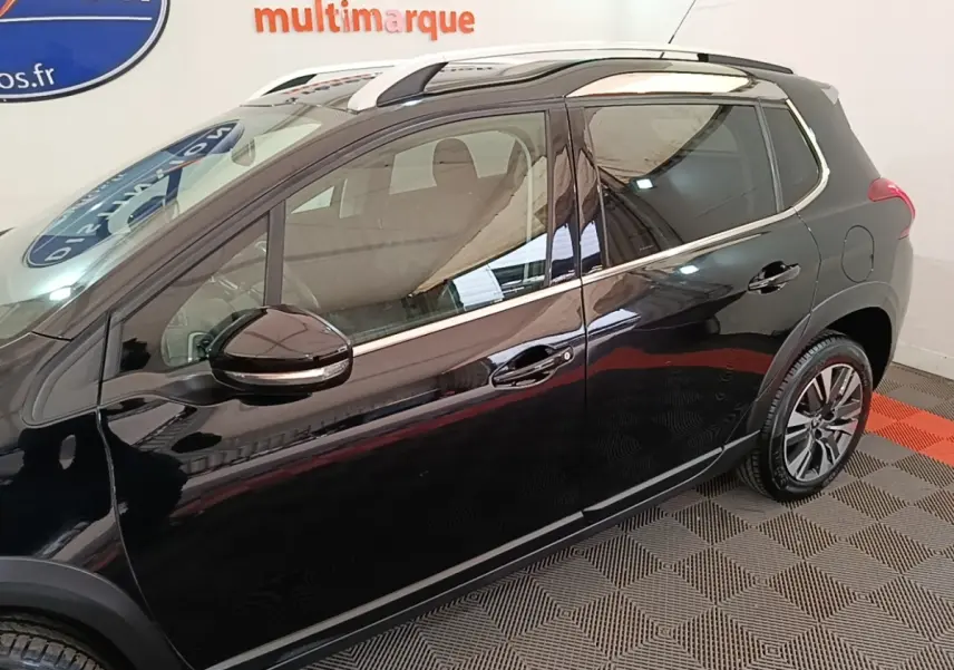 Vue côté gauche d'un Peugeot 2008 noir avec jantes alliage et barres de toit chromées en intérieur showroom.
