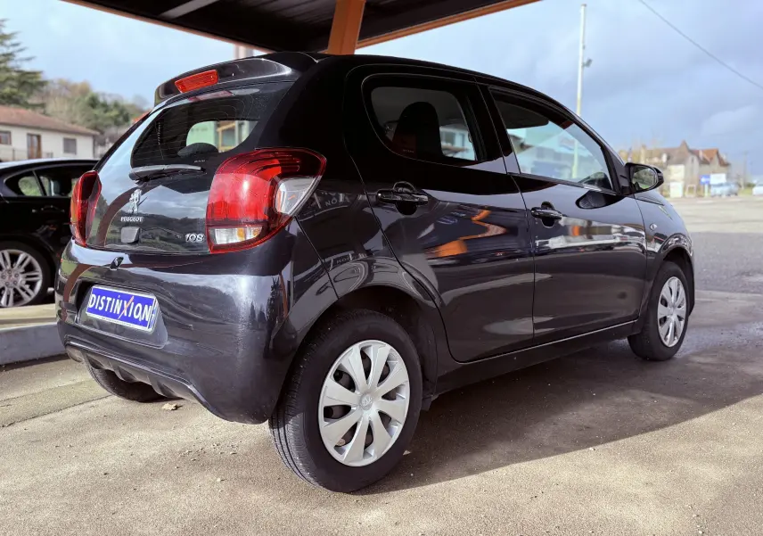 Vue 3/4 arrière droite d'une Peugeot 108 noire 5 portes stationnée sous abri avec jantes en plastique gris clair.