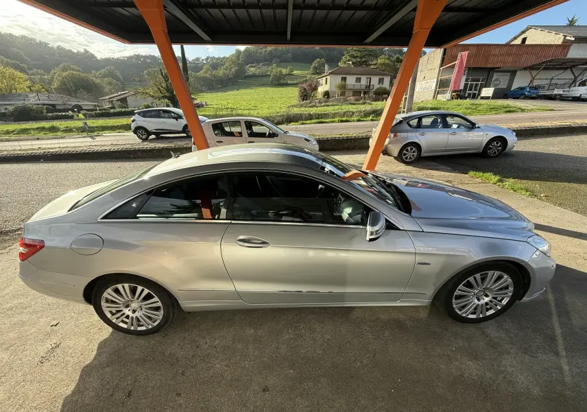 Profil côté gauche d'une Mercedes Classe E 250 Coupé gris métallisé sous un abri orange, avec jantes alliage visibles.