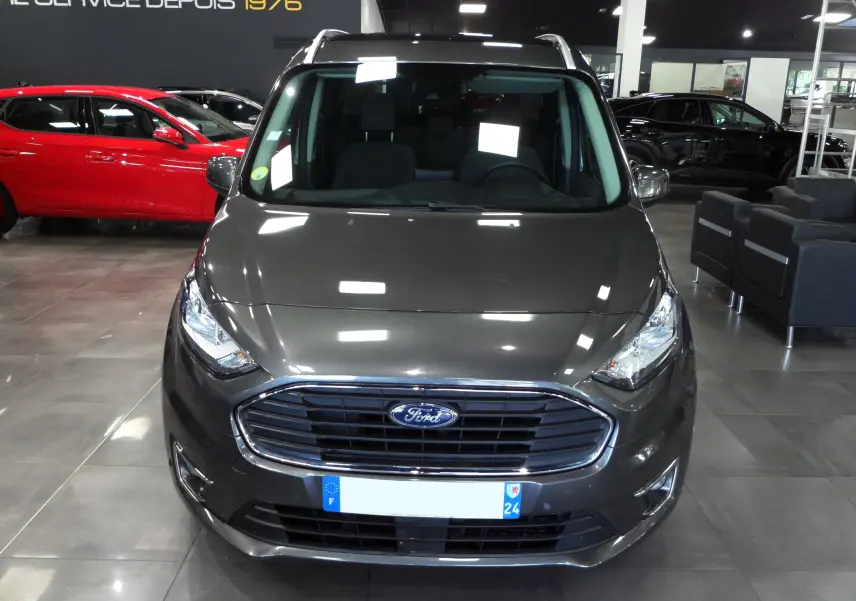 Vue frontale d'un Ford Grand Tourneo Connect gris Magnetic exposé en showroom avec calandre chromée et phares allumés.