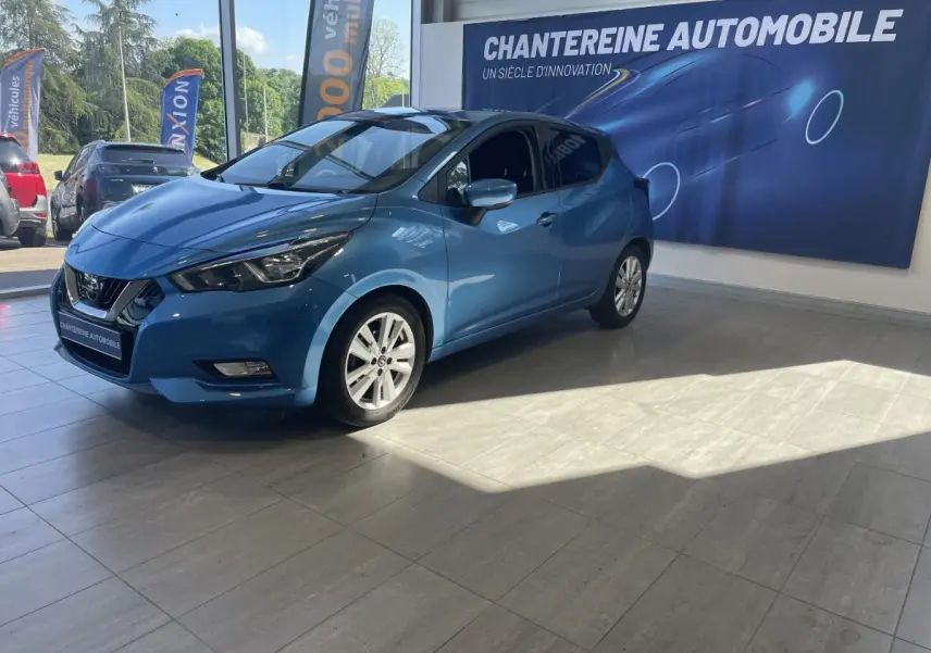 Nissan Micra IG-T 100 N-Connecta bleu vue 3/4 avant droit en showroom avec éclairage naturel et logo concession visible.