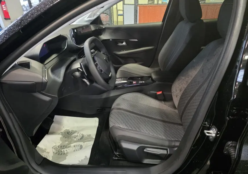 Vue intérieure côté conducteur de la Peugeot 208 noire métallisée, avec sièges tissu gris et tableau de bord moderne.