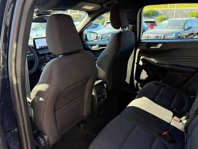 Vue intérieure arrière droite du Ford Kuga 2024 noir Agate, sièges sport noirs avec surpiqûres rouges et tableau de bord visible.