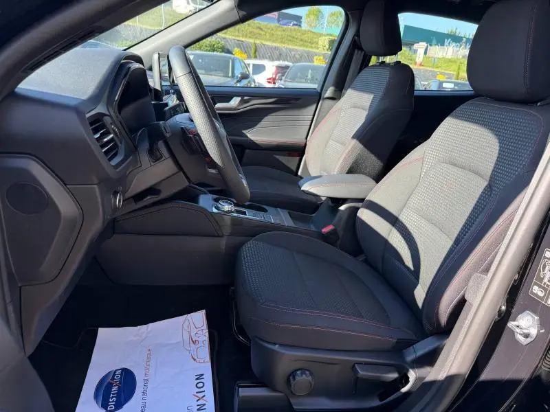 Intérieur avant du Ford Kuga 2.0 EcoBlue 2024, sièges sport tissu noir avec surpiqûres rouges, volant cuir multifonction.