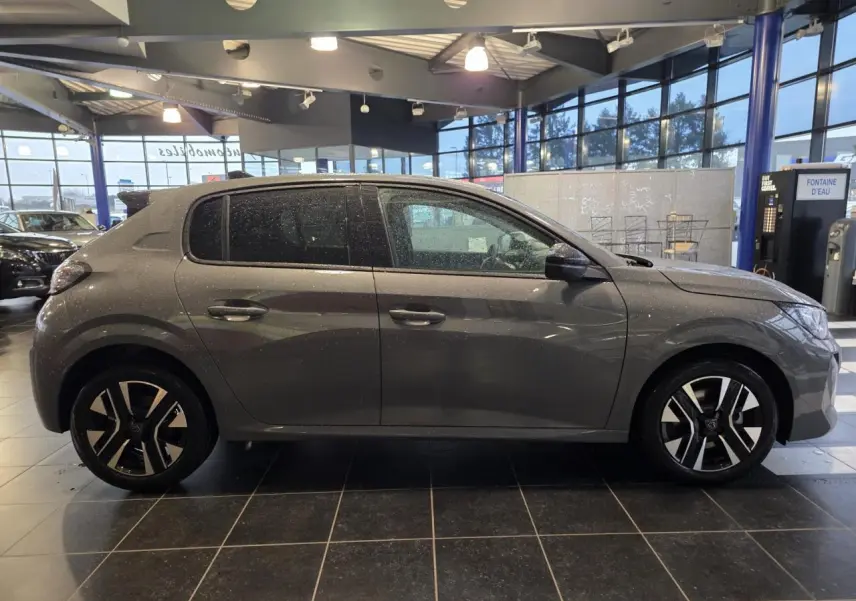 Profil droit d'une Peugeot 208 hybride 2025 en gris selenium métallisé avec jantes alliage bi-ton dans un showroom.