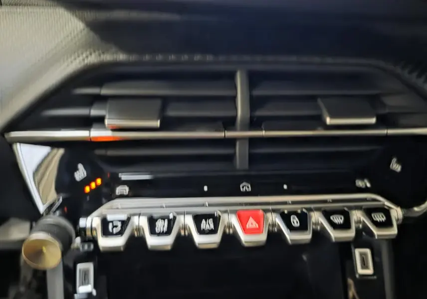 Détail de la console centrale noire brillante de la Peugeot 208 1.2i Hybrid avec boutons argentés et bouton d’alerte rouge.