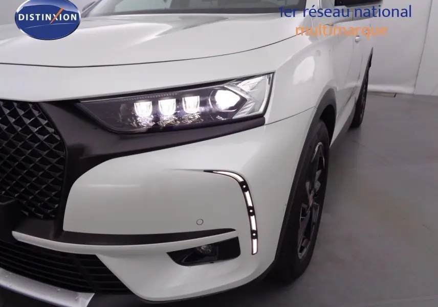 Gros plan sur l'avant droit du DS7 Crossback blanc nacré, mettant en valeur le phare LED allumé et la calandre noire texturée.