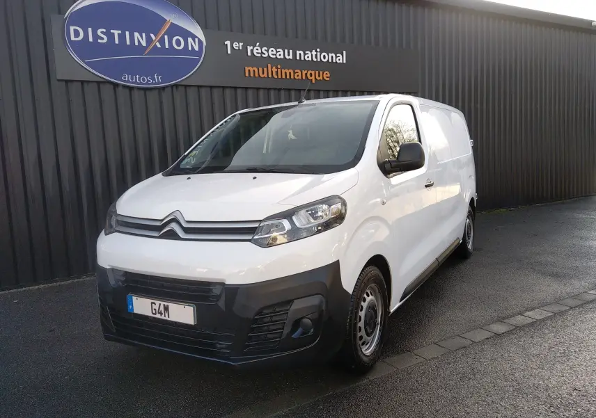 Fourgon Citroën Jumpy blanc en 3/4 avant droit, avec pare-chocs noir et jantes acier, stationné devant un mur gris.