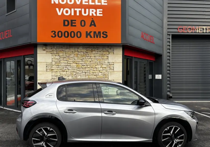 Vue de profil côté gauche d'une Peugeot 208 Hybrid 110 e-DCS6 GT gris clair garée devant un bâtiment moderne.
