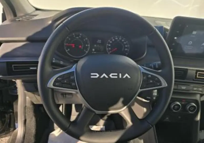 Vue centrée sur le volant noir Dacia avec commandes intégrées, tableau de bord et écran tactile en arrière-plan.