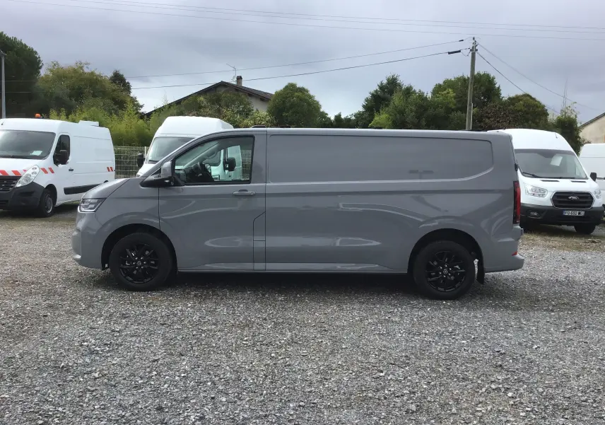 Profil côté gauche d'un Volkswagen Transporter T7 gris stone avec jantes noires sur un parking extérieur.