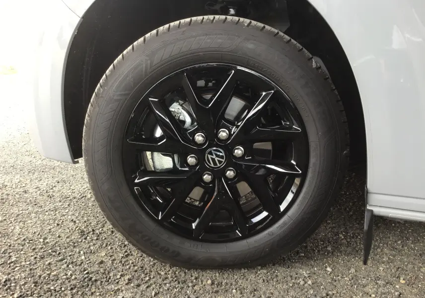 Gros plan sur la roue avant gauche du Volkswagen Transporter gris Stone avec jante noire brillante et logo VW central.