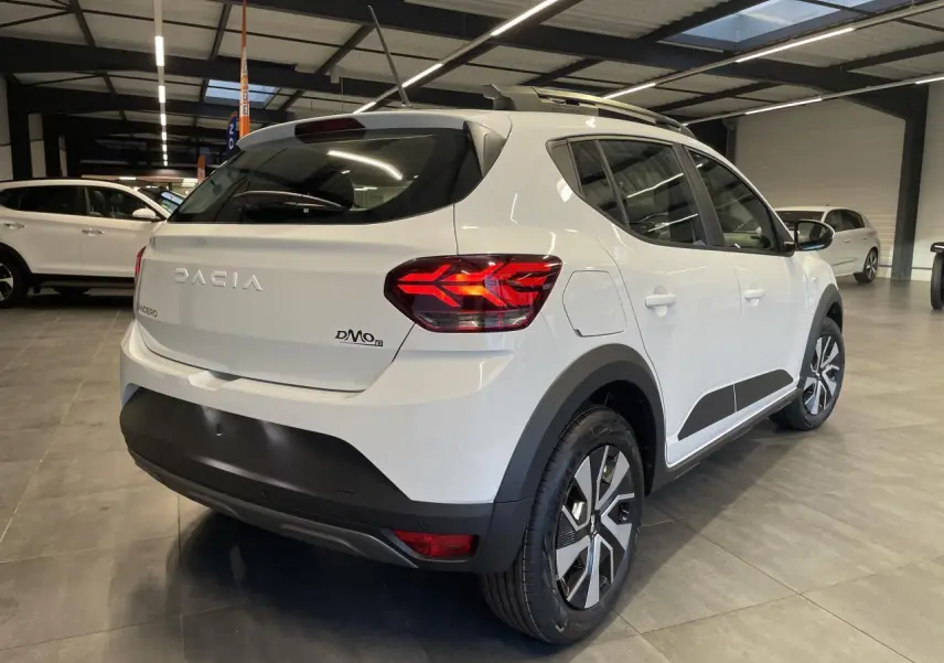 Vue 3/4 arrière droite d'une Dacia Sandero Stepway blanc glacier 2025 avec roue de secours en intérieur showroom.