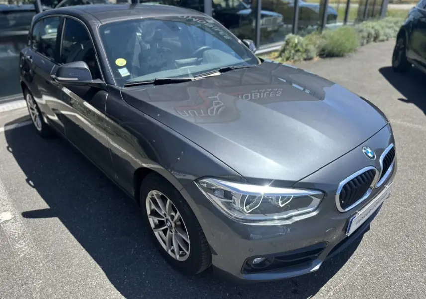 BMW Série 1 F20 gris foncé vue 3/4 avant droit, avec phares LED et calandre double haricot chromée.