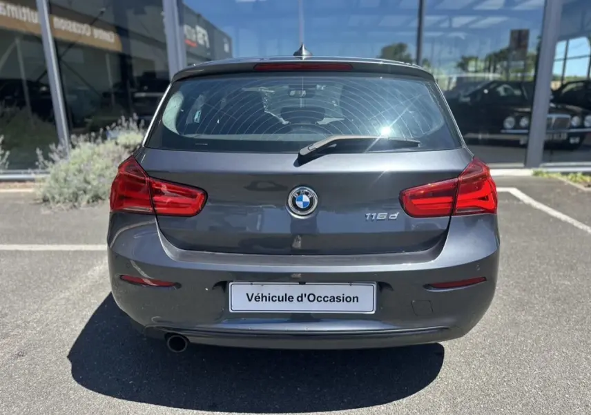 Vue arrière d'une BMW Série 1 F20 gris foncé avec logo et badge 116d, stationnée en extérieur devant un showroom.