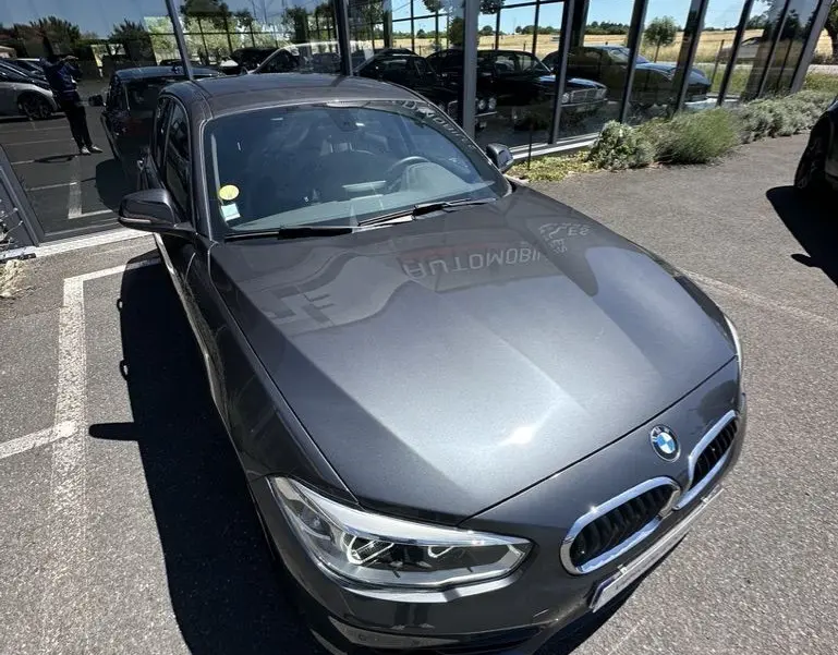 BMW Série 1 F20 gris foncé vue en 3/4 avant droit, stationnée devant une vitrine réfléchissante.