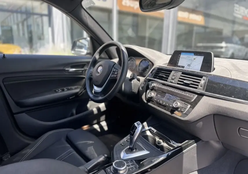 Vue intérieure côté conducteur de la BMW Série 1 F20 gris foncé, avec tableau de bord et écran de navigation visible.