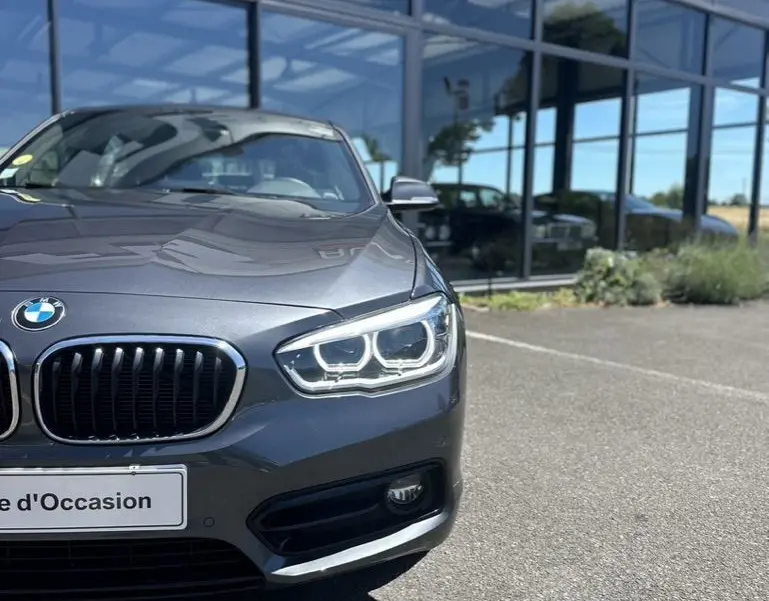 Vue avant droite d'une BMW Série 1 F20 gris foncé avec phares LED allumés devant un showroom.