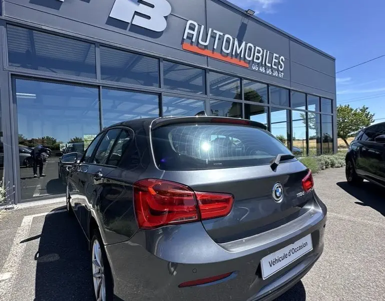 BMW Série 1 F20 gris foncé vue 3/4 arrière droit, avec feu arrière rouge et plaque "Véhicule d'Occasion" visible.