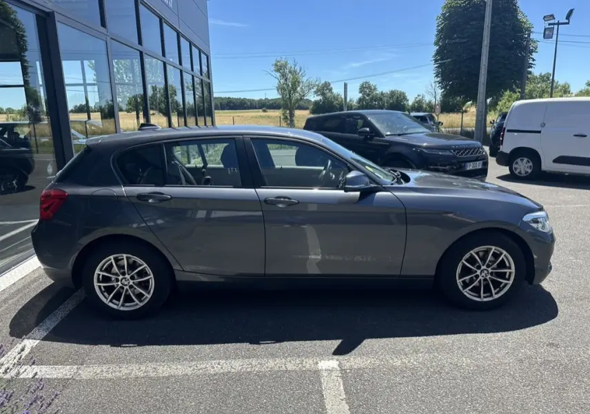 Vue de profil côté gauche d'une BMW Série 1 F20 gris foncé garée devant un bâtiment vitré par temps ensoleillé.